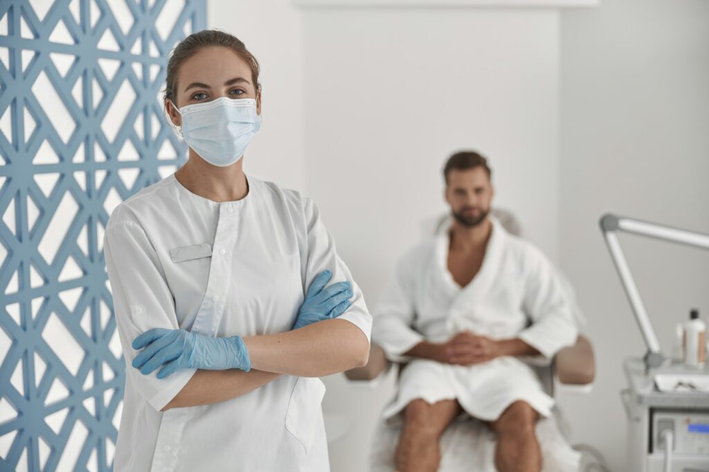 Portrait of professional beautician in protective mask and gloves near patient in cosmetologycal